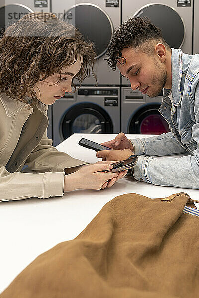 Young couple using mobile phones leaning on table at laundromat