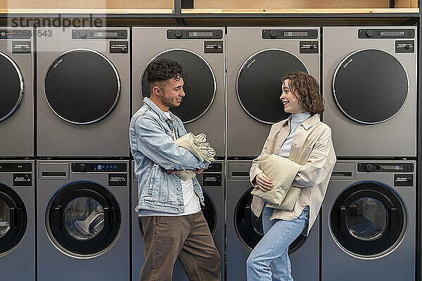 Smiling young friends with sheets leaning on machines at laundromat