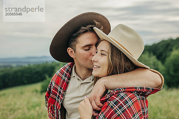 Young man kissing girlfriend wearing plaid shawl on meadow