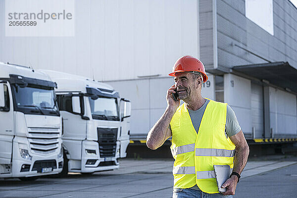 Confident manager wearing protective workwear talking on smart phone at warehouse