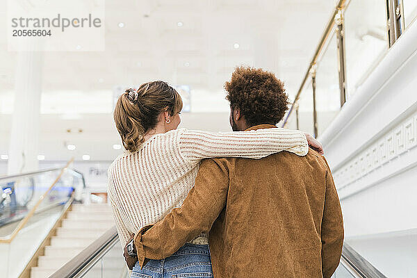 Multiracial couple with arms around standing together on escalator at illuminated shopping mall