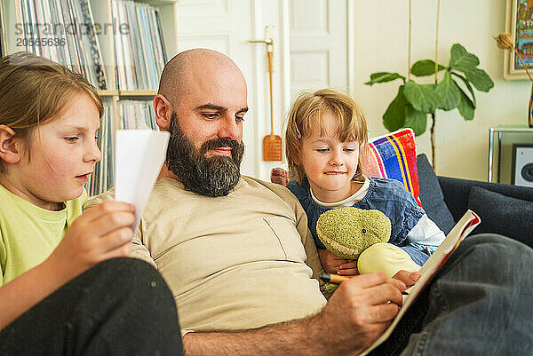 Father writing in book sitting with daughters on sofa at home