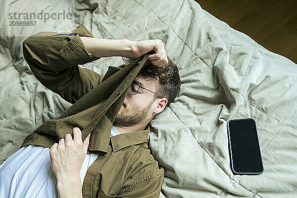 Tired young man lying by smart phone on bed at home