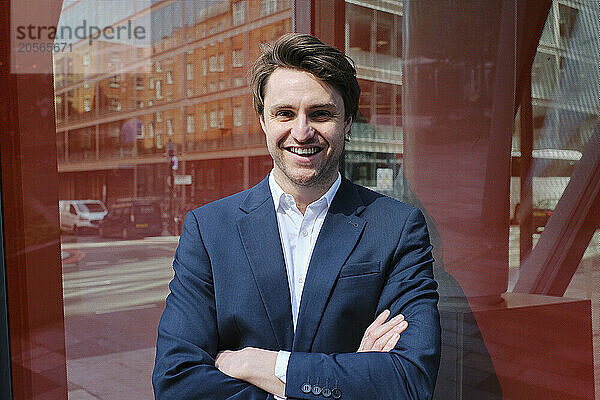 Smiling businessman standing with arms crossed and leaning on red glass