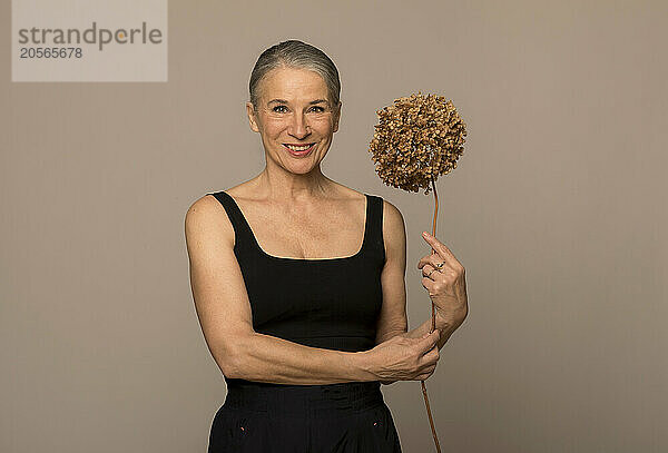 Smiling senior woman holding withered flower in front of brown background