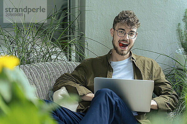 Handsome young man eating cherry tomato sitting with laptop in balcony