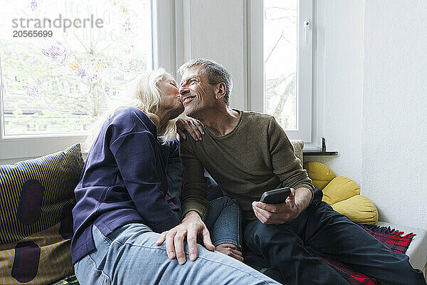 Senior woman kissing man sitting with smart phone at home