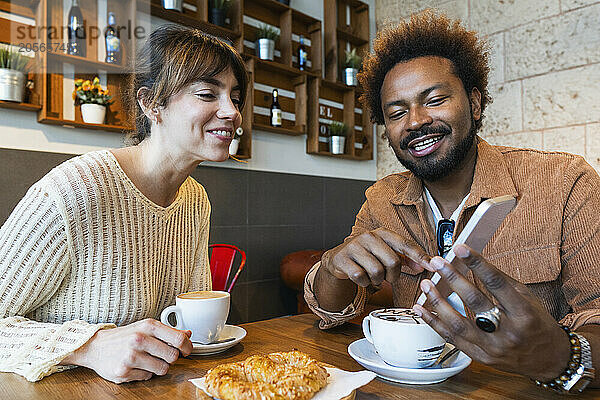 Smiling man sharing smart phone to girlfriend sitting in cafe