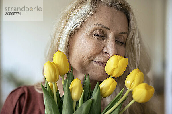 Blond senior woman with eyes closed smelling fresh yellow tulip flowers at home