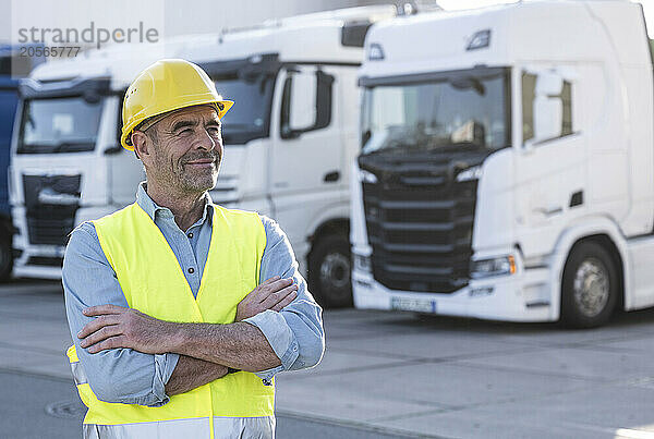 Smiling mature manager with arms crossed looking away standing near trucks