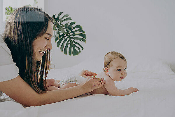 Happy mother massaging baby boy lying on front in bed at home