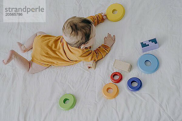 Cute baby boy in orange bodysuit plating with toys in bed at home