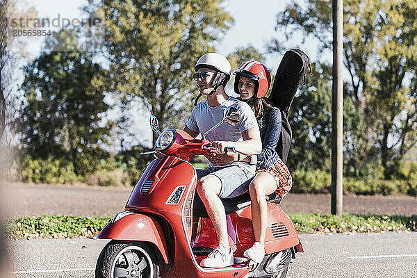 Young woman sitting on motor scooter with boyfriend and guitar bag on back at country road