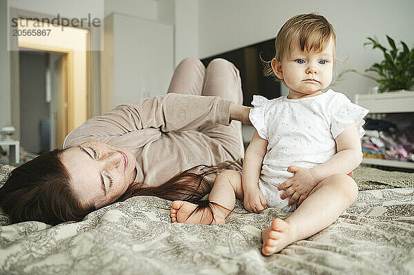 Smiling woman lying by daughter sitting on bed at home