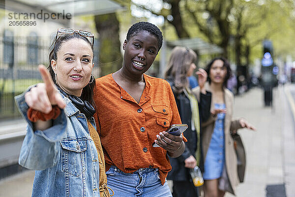 Woman pointing away to friend with smart phone at sidewalk