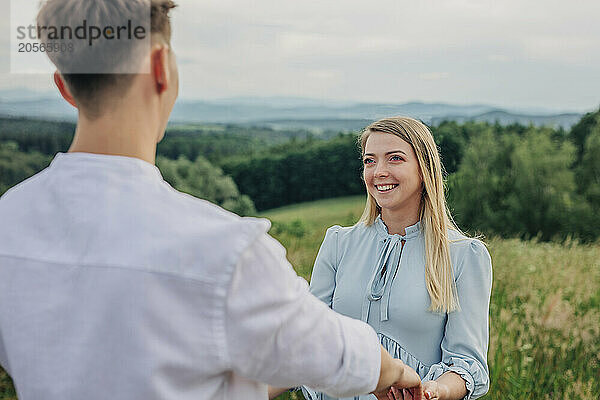 Happy woman holding boyfriends hands and standing in meadow at mountains