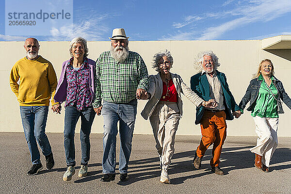 Happy retired men and women holding hands walking in front of wall on sunny day