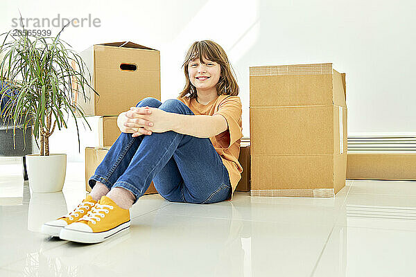 Smiling girl sitting next to boxes and potted plant on floor in new apartment