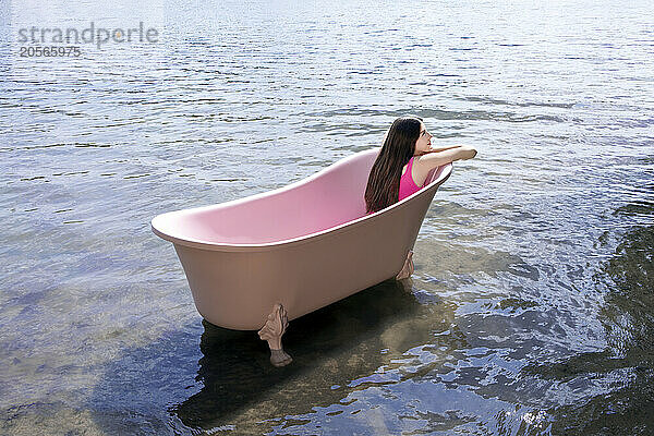 Girl sitting inside pink colored bathtub in lake on sunny day