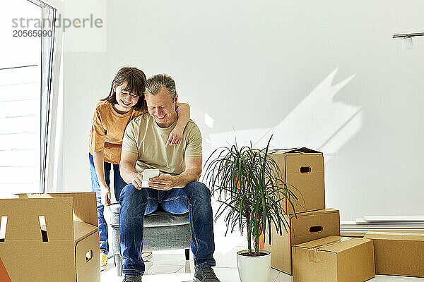 Happy father and daughter using smart phone amidst cardboard boxes in new house