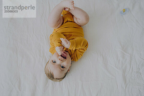 Cute baby boy in orange bodysuit lying on back in bed at home