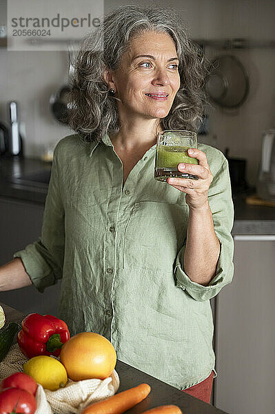 Beautiful gray hair woman holding detox smoothie glass standing in kitchen