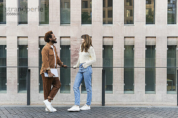 Smiling multiracial couple leaning on railing and talking in front of modern building