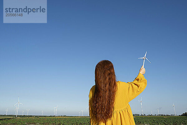 Teenage girl with red hair in a yellow dress holding a model of a wind generator on the background of a blue sky