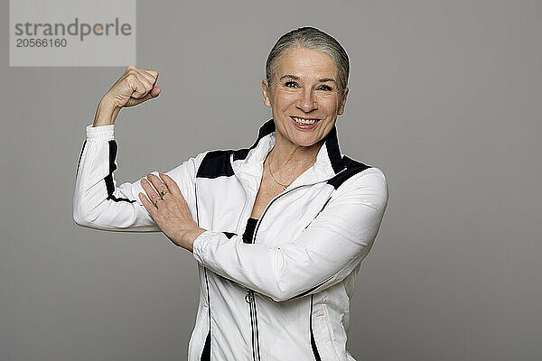 Smiling senior woman flexing muscle against gray background