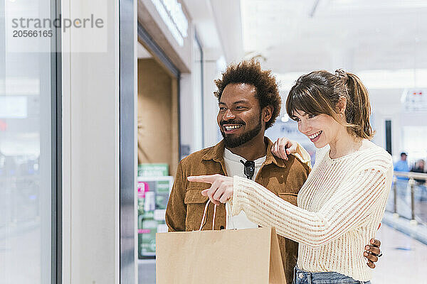 Happy woman with paper bag pointing at window to boyfriend in shopping mall