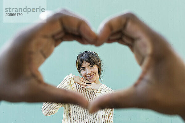 Smiling woman with hand on chin seen through hands of man making heart shape