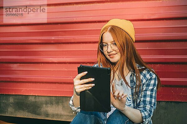 Red haired teenage girl using tablet PC and sitting in front of corrugated iron