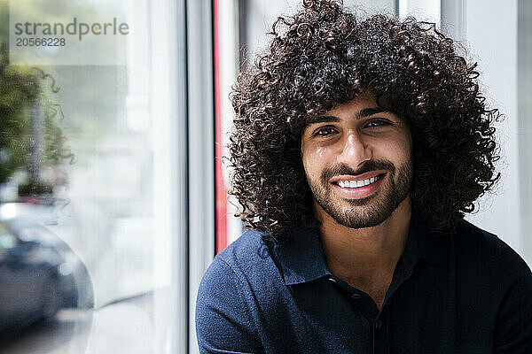 Smiling young handsome businessman with curly hair by window at workplace