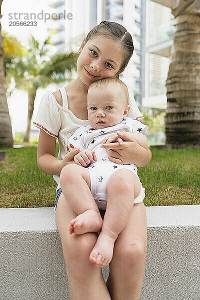 Smiling girl holding baby brother on lap and sitting in back yard