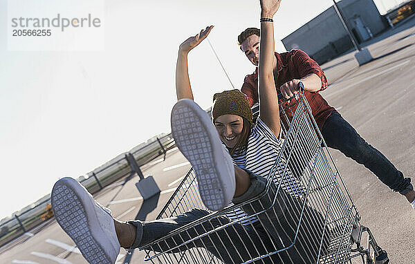 Young man pushing girlfriend sitting in shopping cart under clear sky