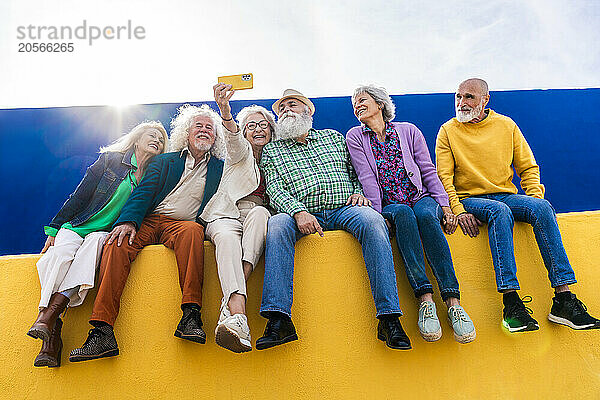 Happy senior friends sitting on yellow wall taking selfie through smart phone