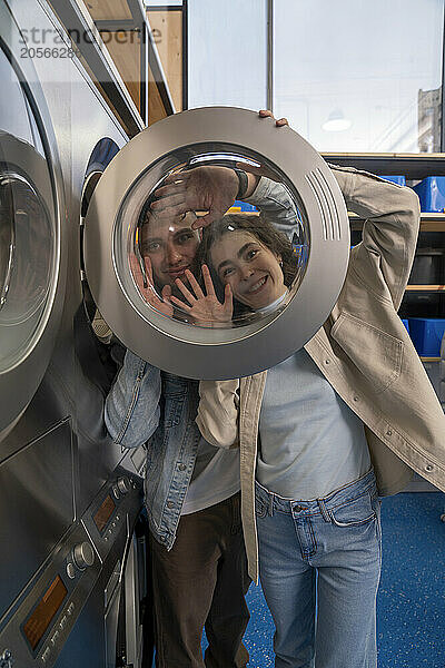 Playful young couple looking through washing machine glass at laundromat