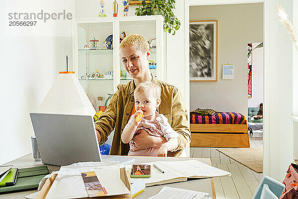 Working mother sitting with toddler daughter and using laptop at home