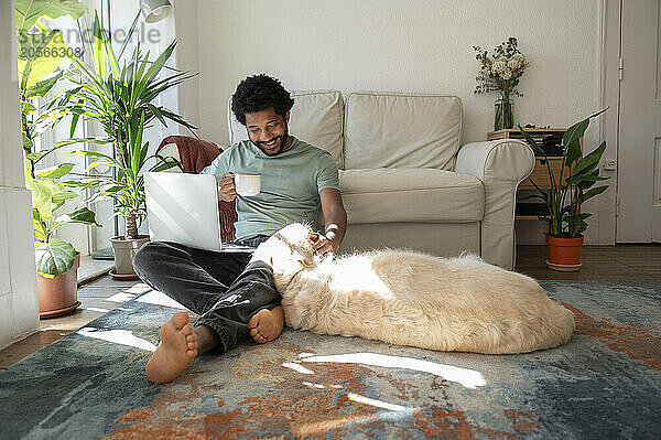 Smiling freelancer holding tea cup and sitting with dog in living room at home