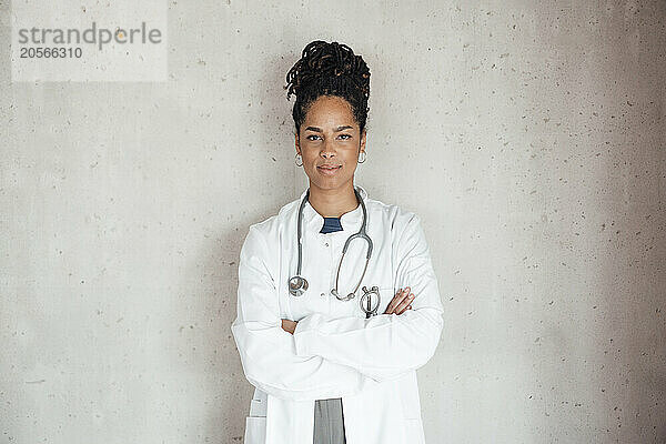 Confident young female doctor wearing lab coat with arms crossed leaning on gray wall at hospital