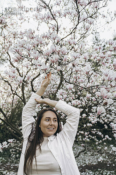 Happy young woman standing with arms raised in front of magnolia tree