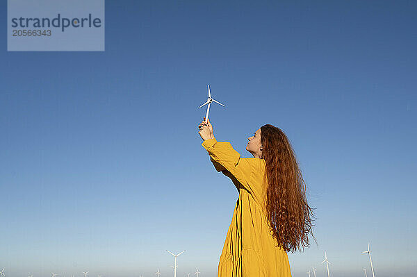 Teenage girl with red hair in a yellow dress holding a model of a wind generator on the background of a blue sky