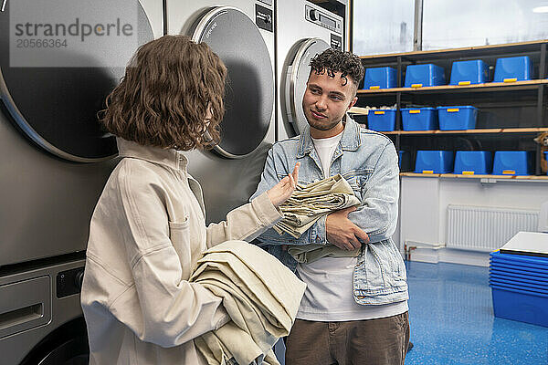 Young friends leaning on machines with sheets talking at laundromat