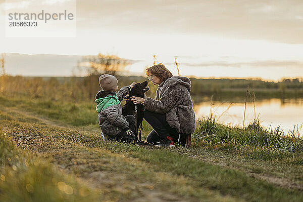 Mother and son crouching with dog on footpath at sunset