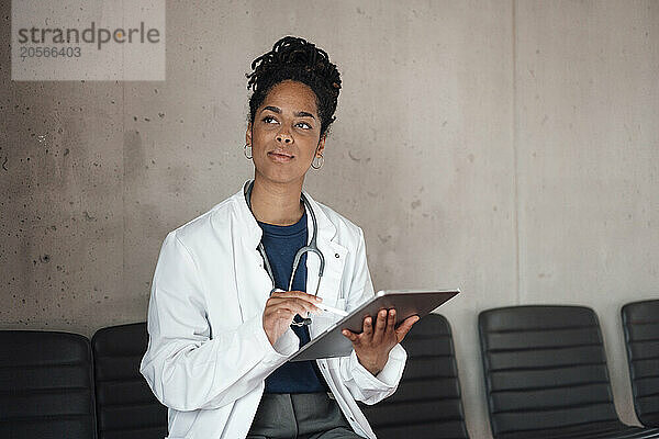 Thoughtful young female doctor with tablet PC sitting on chair in hospital lobby