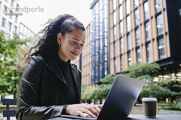 Smiling young businesswoman using laptop sitting with disposable cup at sidewalk cafe