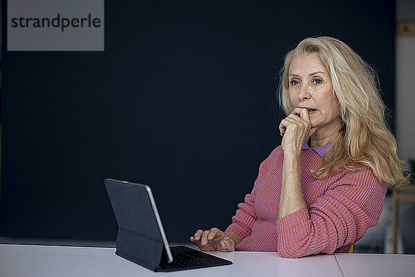 Thoughtful senior woman sitting with tablet PC at table