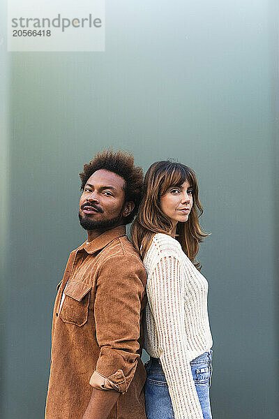 Confident multiracial couple standing back to back against gray background