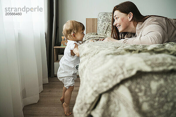 Cheerful mother playing with daughter standing with support of bed at home