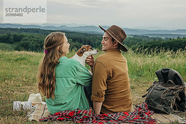 Happy boyfriend and girlfriend playing with dog on meadow at mountain